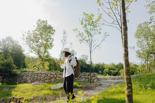 Photo of a young woman hiking with a backpack through a beautiful nature on a sunny summer day, while checking often the maps on her mobile phone