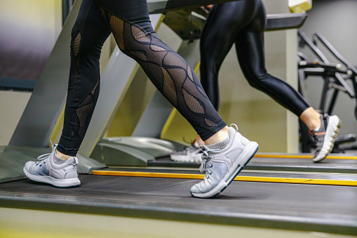 Low section of two women in leggings and sneakers working out on treadmills in the gym, healthy life and habits