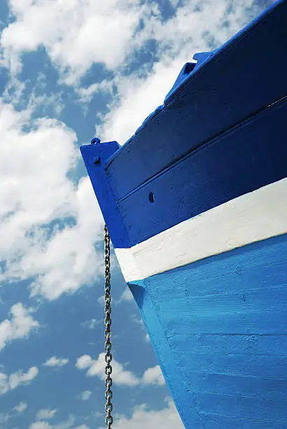 Side view of a wooden white and blue fisherman boat with its chain anchor under a cloudy sky.