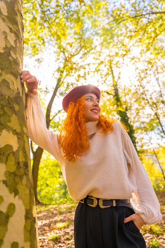 Red-haired woman leaning against a tree in a forest park smiling at sunset