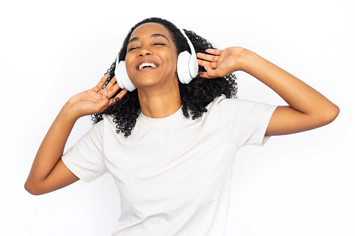 Excited multiracial woman dancing in headphones. Happy young female model with dark curly hair in white T-shirt with closed eyes, laughing, enjoying music. Modern technology, music concept