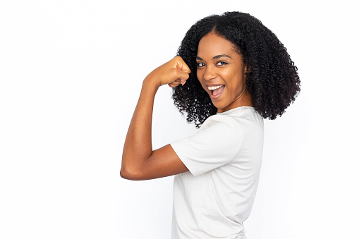 Happy African American woman showing bicep. Strong young female model with dark curly hair in white T-shirt looking at camera, smiling with raised fist, demonstrating power. Success, strength concept