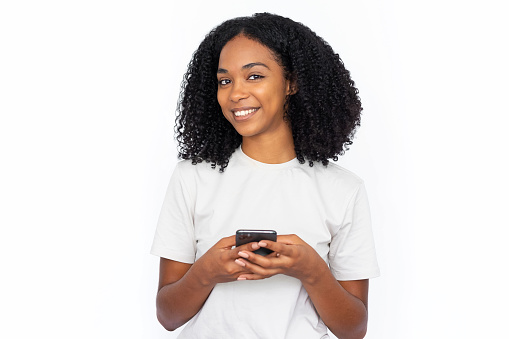 Pleased African American woman using phone. Happy young female model with dark curly hair in white T-shirt looking at camera, smiling, dialing ads number. Modern technology, advertising concept