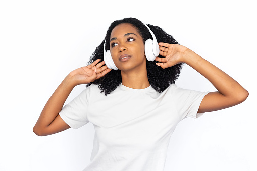 Pleased multiracial woman listening to music. Relaxed young female model with dark curly hair in white T-shirt looking away, dancing, enjoying song in headphones. Modern technology, music concept