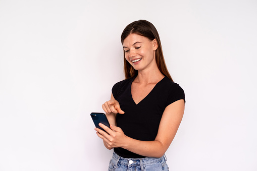 Portrait of happy young woman surfing internet on smartphone over white background. Caucasian lady wearing black T-shirt and jeans texting message on mobile phone. Mobile communication concept