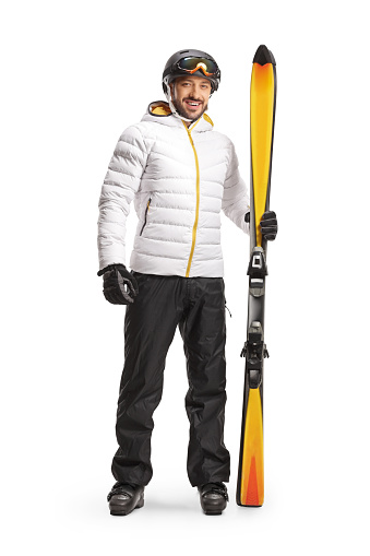 Full length portrait of a male skier with equipment standind and holding a pair of skis isolated on white background