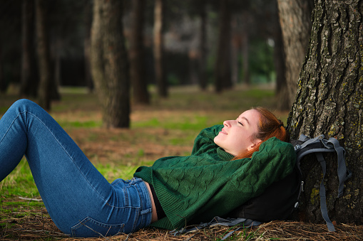 Redhead young woman resting lying under tree in forest. Relaxing in nature.