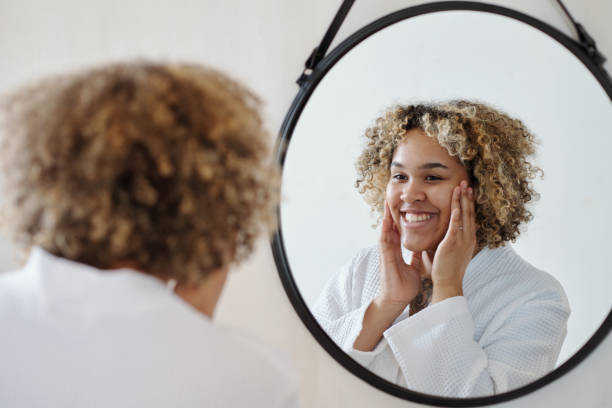 istock Young smiling black woman touching her face while looking in mirror 1448709927