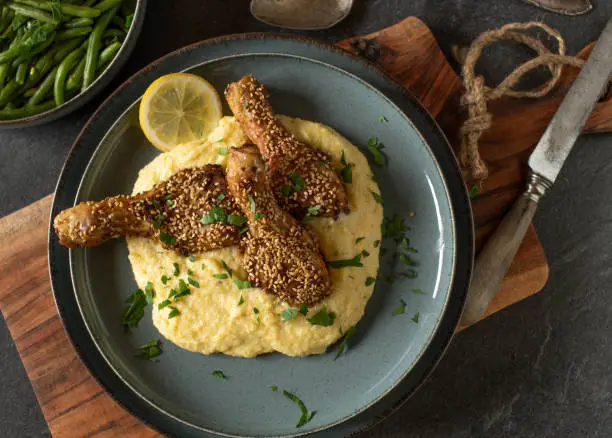 Homemade delicious chicken meal cooked with oven baked drumsticks marinated with harissa paste and topped with sesame seeds. Served with creamy polenta and princess beans on a plate.  Overhead view