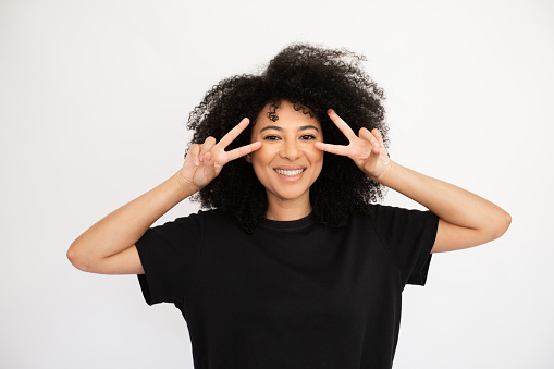 Joyful young woman showing V-signs near her eyes. Hispanic female model with afro hairstyle and brown eyes in black T-shirt smiling and making victory signs near eyes. Happiness, peace concept