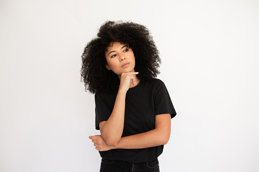 Pensive young woman looking away holding thumb on her chin. Hispanic female model with afro hairstyle and brown eyes in black T-shirt looking away touching chin with thumb. Thought, dream concept