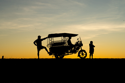 Asian village lifestyle, silhouette of a local Thai male father and son with their Skylab taxi vehicle by a freshwater lake during dawn sunrise time, very beautiful scenic view