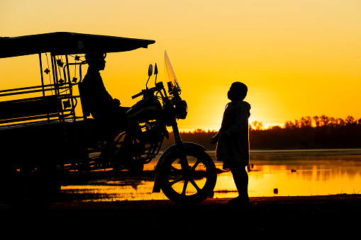 Asian village lifestyle, silhouette of a local Thai male father and son with their Skylab taxi vehicle by a freshwater lake during dawn sunrise time, very beautiful scenic view