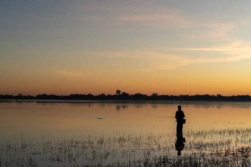 Asian village lifestyle, silhouette of a local Thai male fisherman fishing by a freshwater lake during dawn sunrise time, very beautiful scenic view