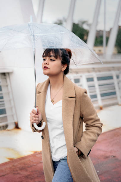 Pretty woman walking in the city during rainy day holding a transparent umbrella. She is wearing makeup and looking at front.