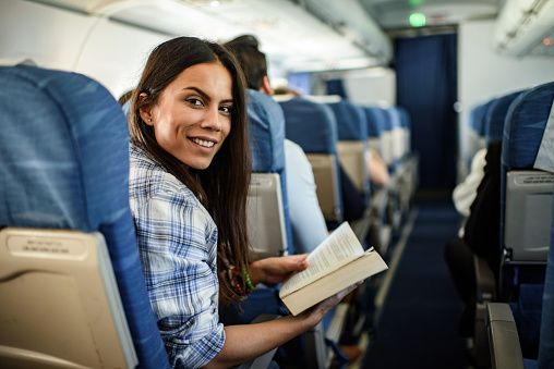 Young happy woman reading a book while traveling by plane and looking at camera.