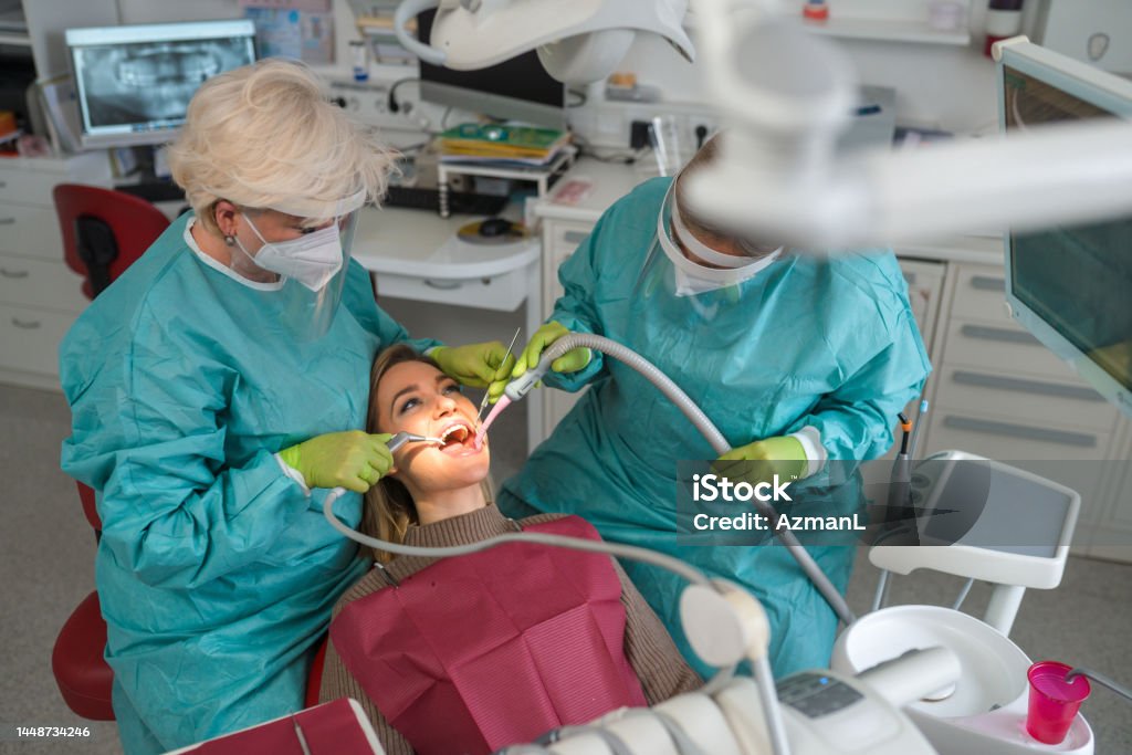 Young Caucasian Woman Having A Dental Check-Up Exam During Pandemic While Caucasian female dentist working with young patient female dental assistant helping around. Both wearing a protective gowns, face masks and gloves during covid-19 pandemic. Dentist Stock Photo