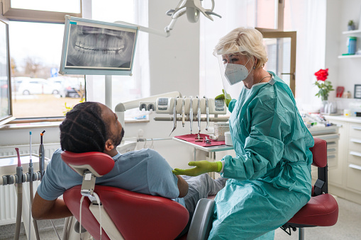 Senior Caucasian female dental professional and radiologist explaining X-ray image to an African-American male patient during covid-19 pandemic.