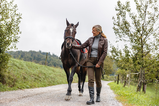A happy adult caucasian woman caressing the horse as they are walking on a country road surrounded by pastures and beautiful nature. The woman is a horse rider and is wearing boots and a leather jacket. The lady is holding the reins and looking at the horse. The weather is cloudy.