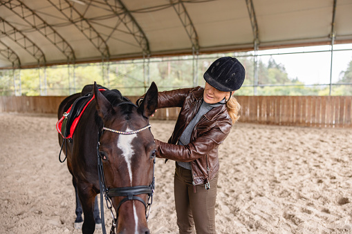 A focused adult caucasian blonde female horse rider taking care of her horse after a riding session in a riding arena. She is grooming her horse with a brusg. The woman is wearing a black riding helmet and a brown leather jacket. The horse is saddled and has a bridle. They are standing on sand.