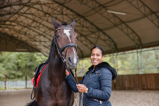 A happy  black young adult female horse trainer in a winter jacket holding the reins while standing next to the horse in a countryside covered riding arena. They are both looking at the camera. The riding arena is surrounded by a wooden fence. The weather is cold and gloomy. The attractive woman is looking at the camera.