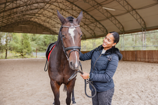 An attractive black young adult female horse rider smiling while petting her brown horse. They ae standing in the midle of the riding arena. The arena in covered and located in the beautiful rural area. The female is wearing a winter jacket since it is a cold fall day. The woman looks happy while connecting with her horse.