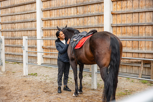A beautiful black young adult female horse owner caressing and hugging a brown horse in front of the stable. The female is smiling while standing next to her horse. They are surrounded by a fence. The weather is cold and cloudy. The black female is wearing a winter jacket and some jeans. The horse has a saddle on.