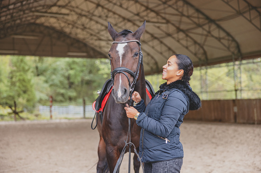 A smiling black young adult woman having a good time while spending time with a horse. They are standing on a sand in a countryside riding arena. She is smiling while looking at the horse. The woman is holding the reins and looks relaxed. She is dressed in a winter jacket since it is cold and cloudy.