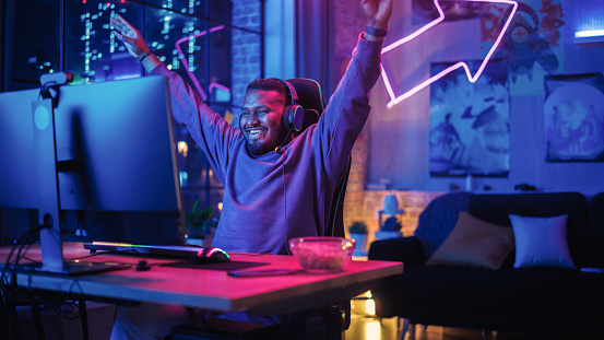 Juegos en casa: Jugador negro jugando videojuegos en línea en la computadora personal. Elegante jugador masculino afroamericano disfrutando del torneo en línea en su apartamento Loft. photo
