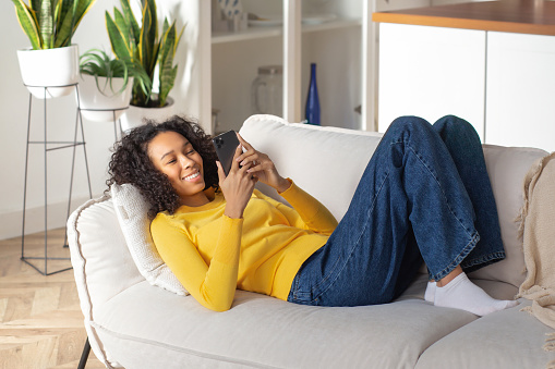 Young African American woman uses a mobile phone to communicate in a social media. Beautiful happy female shopping online lying on the couch. Relax at home, weekend concept