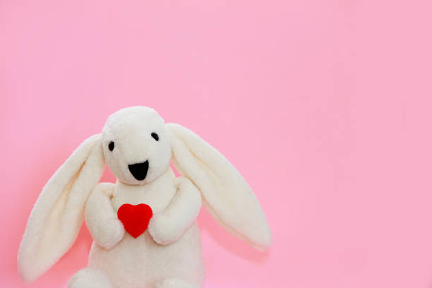A white fluffy rabbit holds a red heart in its paws on  pink background