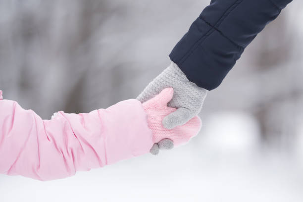 Little daughter and young adult mother holding each other hands in warm gloves on nature background. Walking and spending time together in cold winter day. Closeup. Back view. stock photo