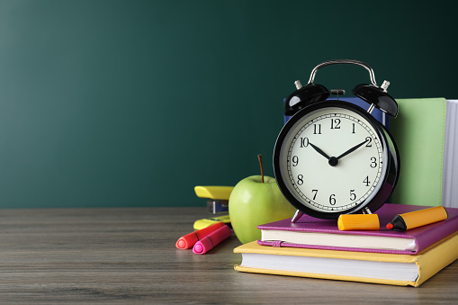 Alarm clock and different stationery on wooden table near green chalkboard, space for text. School time