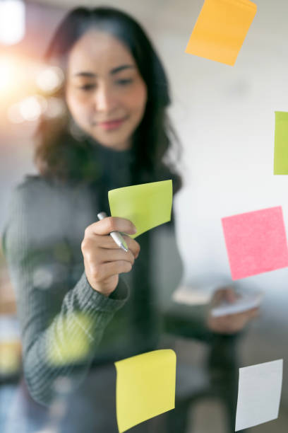 istock Young smiley attractive,businesswoman using post it notes in glass wall to writing strategy business plan to development grow to success 1448763658