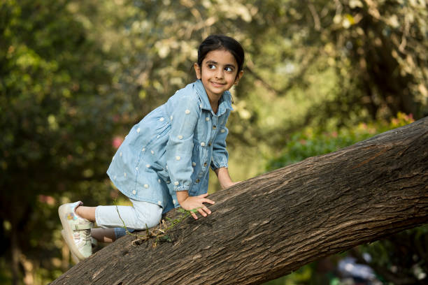 Playful elementary Indian girl laughing and climbing on tree trunk while enjoying weekend in park