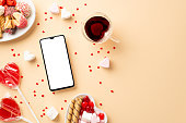istock Valentine's Day concept. Top view photo of smartphone plates with sweets cookies candies lollipops and heart shaped glass cup of drinking on isolated beige background with blank space 1448766286
