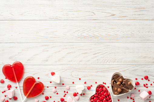 Valentine's Day concept. Top view photo of heart shaped saucers with confectionery chocolate candies and lollipops on white wooden table background with blank space