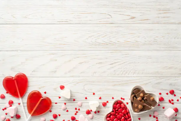 Photo of Valentine's Day concept. Top view photo of heart shaped saucers with confectionery chocolate candies and lollipops on white wooden table background with blank space