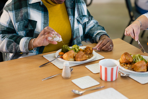 A close-up shot of unrecognisable people visiting a warm hub/food bank which is a safe place for people to enjoy a warm and friendly environment in the community with the current cost of living increasing. An unrecognisable young woman is sitting at a table with a senior woman, they are eating a roast dinner and the young woman is sprinkling salt.