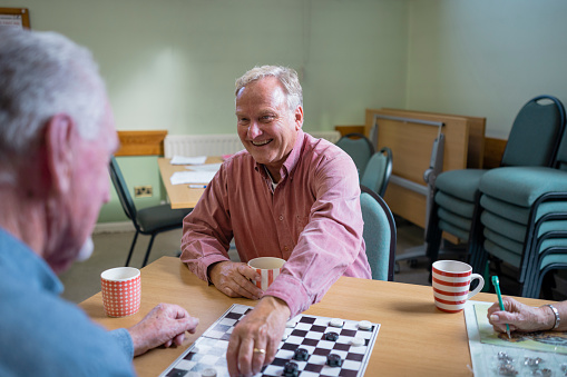 An over the shoulder shot of a small group of people visiting a warm hub/food bank which is a safe place for people to enjoy a warm and friendly environment in the community with the current cost of living increasing. Two senior friends are sitting around a table, they are playing a game of checkers.