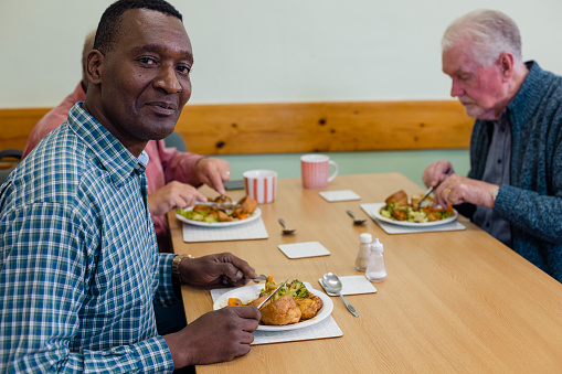 A shot of people visiting a warm hub/food bank which is a safe place for people to enjoy a warm and friendly environment in the community with the current cost of living increasing. Three senior men are sitting at a table and one man is smiling and looking at the camera, they are eating a roast dinner.