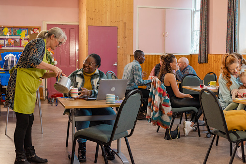 A shot of a medium group of people visiting a warm hub/food bank which is a safe place for people to enjoy a warm and friendly environment in the community with the current cost of living increasing. One young woman is sitting at a table, she is using a laptop to study while a volunteer pours her a cup of tea.