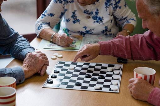 A close-up shot of an unrecognisable small group of people visiting a warm hub/food bank which is a safe place for people to enjoy a warm and friendly environment in the community with the current cost of living increasing. Three senior friends are sitting around a table, they are playing a game of checkers.
