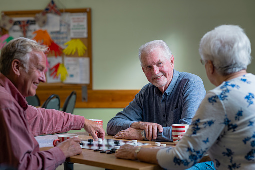 A shot of a small group of people visiting a warm hub/food bank which is a safe place for people to enjoy a warm and friendly environment in the community with the current cost of living increasing. Three senior friends are sitting around a table, they are playing a game of checkers.