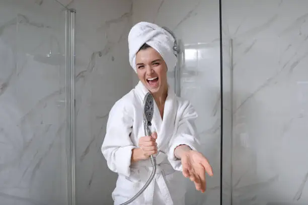 Beautiful young woman singing into showerhead in bathroom