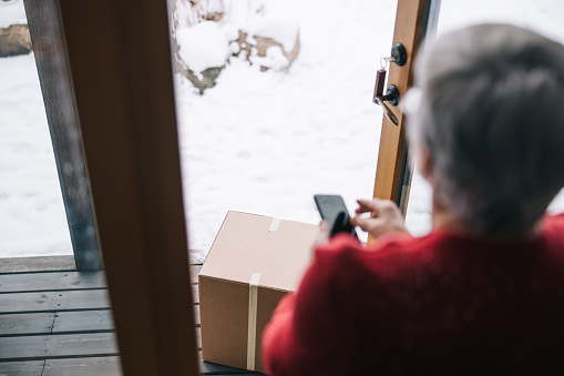 Senior woman using smartphone, while receiving a package at home during Christmas times.