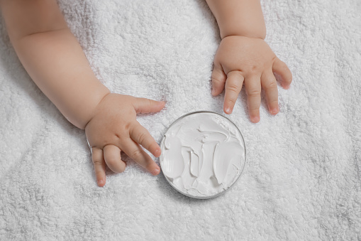 Cute little baby playing with jar of moisturizing cream on towel, closeup
