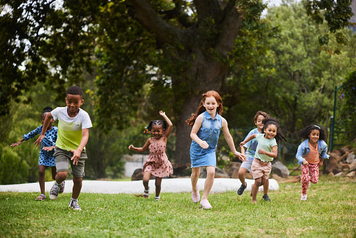 Diverse group of laughing young children running together on grass while playing in a park in summer