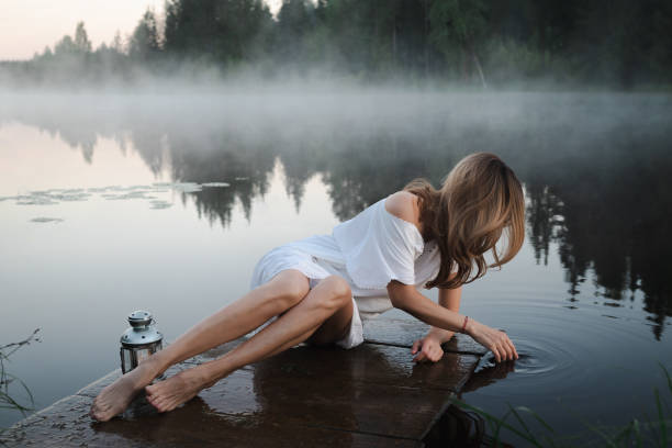 A barefoot middle-aged adult woman in a white dress with a decorative lantern in her hands against the background of a morning lake with fog. The concept of mysticism.