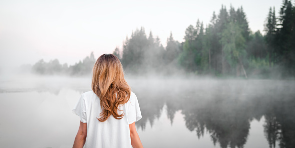 A barefoot middle-aged adult woman in a white dress with a decorative lantern in her hands against the background of a morning lake with fog. The concept of mysticism.